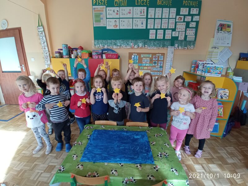 Na zdjęciu dzieci prezentują samodzielnie wycięte wyklejone wydzieranymi kawałkami żółtej kartki gwiazdy do zrobienia flagi Unii Europejskiej.
