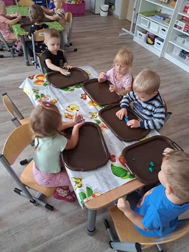 Zdjęcie przedstawia dzieci siedzące przy stolikach  które na plastikowych  brązowych tackach manipulują kolorową plasteliną.