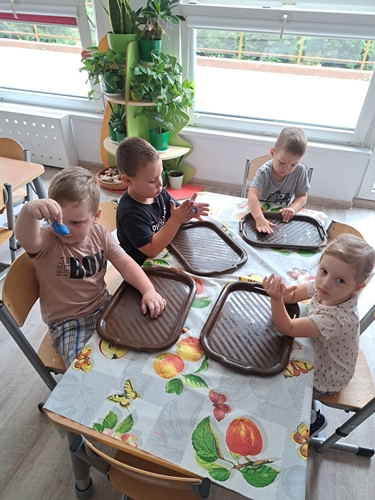Zdjęcie przedstawia dzieci siedzące przy stolikach  które na plastikowych  brązowych tackach manipulują kolorową plasteliną.