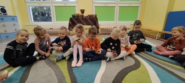 Zdjęcie drugie przedstawia grupę dzieci siedzącą w rzędzie na dywanie. Dzieci trzymają w rękach instrumenty przypominające żaby. Dzieci próbują na nich zagrać.