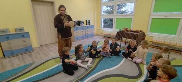 Zdjęcie trzecie przedstawia grupę dzieci siedzącą w półkolu na dywanie. Dzieci trzymają w dłoniach instrumenty i próbują na nich zagrać. Za plecami dzieci stoi mężczyzna grający na bębnie.