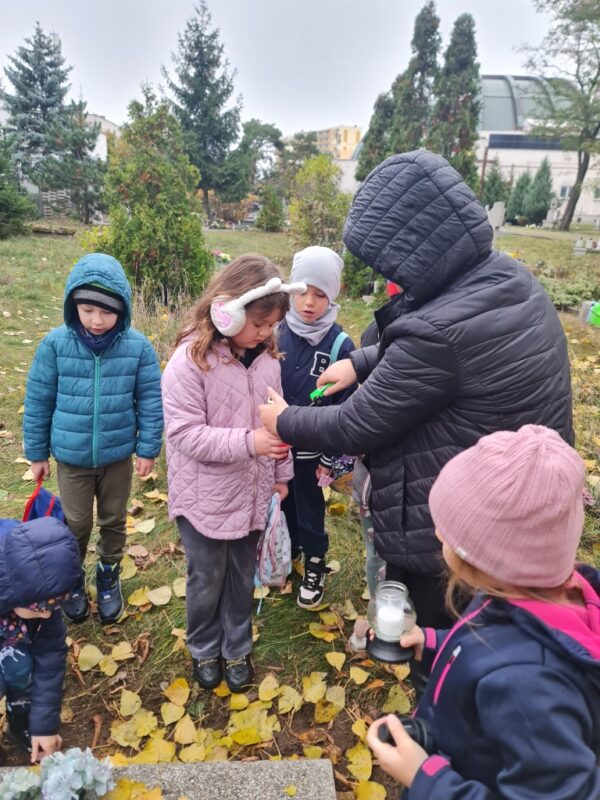Dzieci wraz z nauczycielem zapalają znicze na opuszczonych grobach.