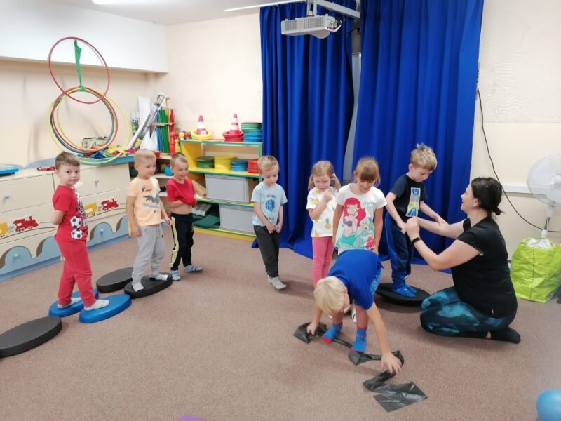 Dzieci ustawione w rzędzie wykonują różne zadania sportowe: rozciąganie na taśmie, utrzymanie równowagi na kole balansującym.