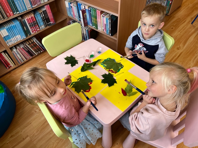 Troje dzieci siedzi przy stoliku, malują farbami liście i odbijają je na kartce.