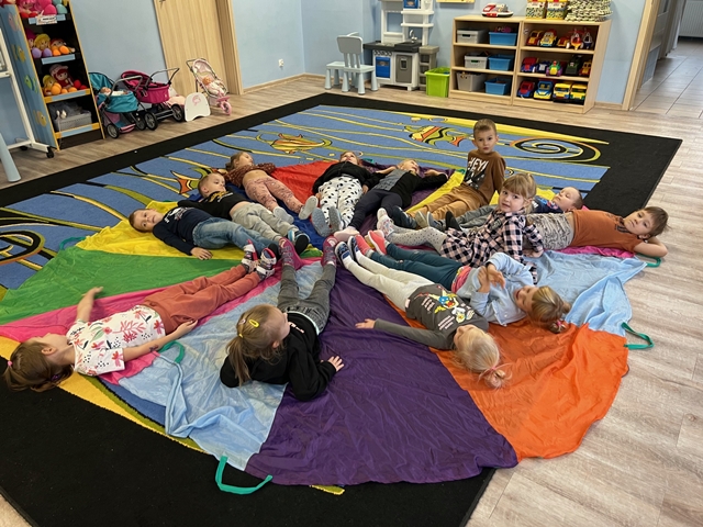 Dzieci leżą na kolorowej chuście animacjnej ułożonej na dywanie. Nogi mają skierowane do środka.