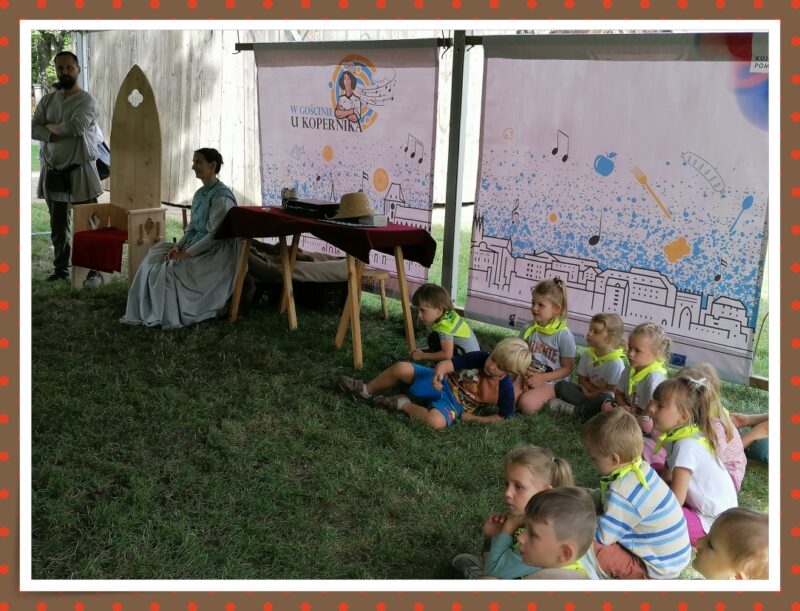 Dzieci leżąc na trawce słuchają prelekcji o Mikołaju Koperniku.