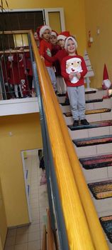 Zdjęcie pierwsze przedstawia grupę dzieci ustawioną w rzędzie. Na pierwszym planie widać chłopca ubranego w czerwoną koszulkę ze świętym Mikołajem i mikołajkową czapkę. Dzieci znajdują się na schodach na korytarzu przedszkola.