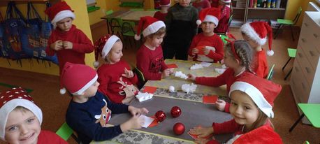 Zdjęcie drugie przedstawia grupę dzieci ubraną w czerwone koszulki i mikołajkowe czapki. Dzieci siedzą wokół stołu i wykonują zadanie.