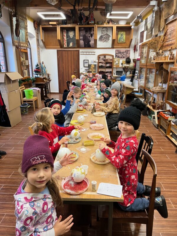 Dzieci siedzą przy długim stole i ozdabiaja bombki przy użyciu kleju i brokatów. Dzieci na głowie mają czapki.