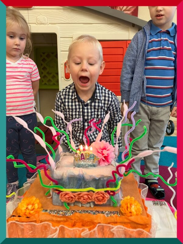 Na zdjęciu chłopiec próbuje zdmuchnąć świeczki z tortu wykonanego z pudełek, kreatywnych drucików, sztucznych kwiatów itp. przez nauczyciela grupy.