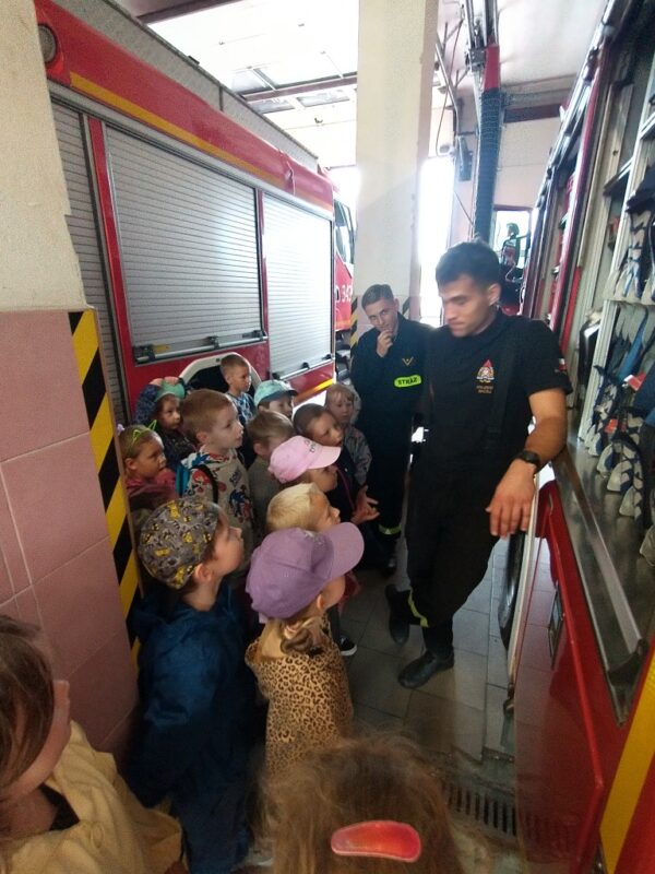 Na zdjęciu grupa 6-latków stoi naprzeciwko wozu strażackiego i z uwagą śledzi prezentację jednego ze Strażaków, związaną z jego wyposażeniem. Dzieci zgromadzone są w garażu jednostki gaśniczo-pożarniczej pomiędzy dwoma wozami, gotowymi do akcji.