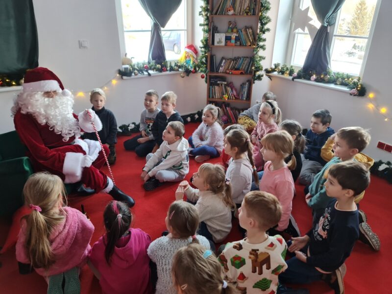 Grupa dzieci siedzi na dywanie w pokoju Świętego Mikołaja i rozmawia z nim, Mikołaj siedzi na zielonym fotelu, trzyma laskę, pyta dzieci o wymarzone prezenty.
