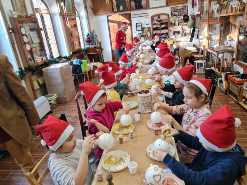 Dzieci są pracowni warsztatowej, ozdabiają bombki świąteczne przy pomocy kleju i brokatu, siedzą wspólnie przy długim stole.