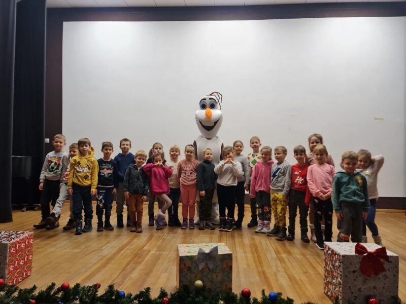 Grupa dzieci stoi obok siebie na scenie z Olafem - postacią z bajki "Kraina lodu" i pozuje wspólnie do zdjęcia.