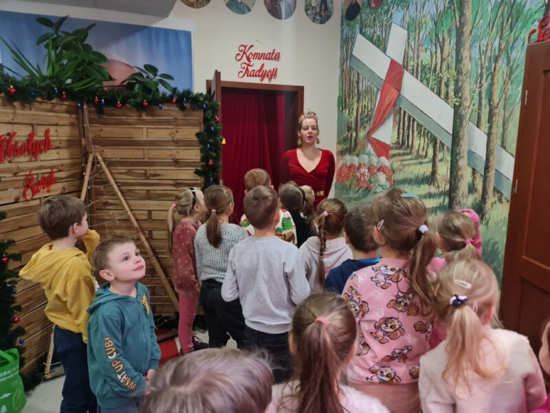 Dzieci słuchają opowieści prowadzącej o Komnacie tradycji, prowadząca podaje instrukcje jak się do niej dostać, pokazuje mapę, jest ubrana w długą czerwoną suknię, dzieci słuchają z zaciekawieniem.