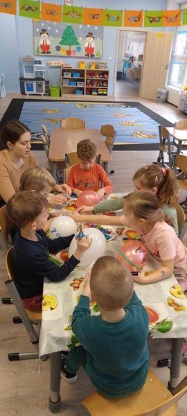  Zdjęcie drugie przedstawia grupę dzieci siedzącą przy stolikach i ozdabiającą balony. W lewym górnym rogu znajduje się pani, pomagająca dzieciom w wykonaniu zadania.
