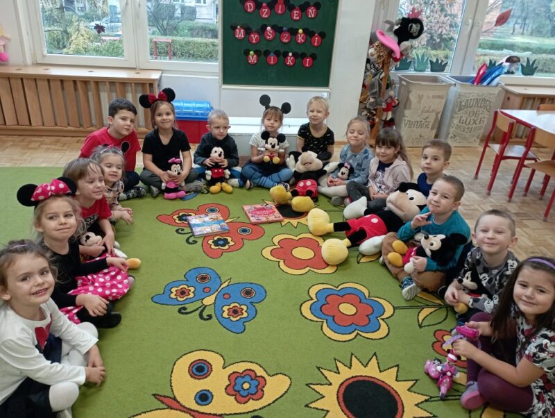 Na zdjęciu dzieci siedzą w półkole, niektóre przebrane za myszkę miki, trzymają przytulanki z bohaterką dnia.