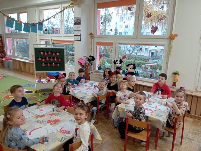 Na zdjęciu cała grupa dzieci przy stolikach wykonuje pracę plastyczną - wykleja z czerwonego papieru twarz Myszki Miki.