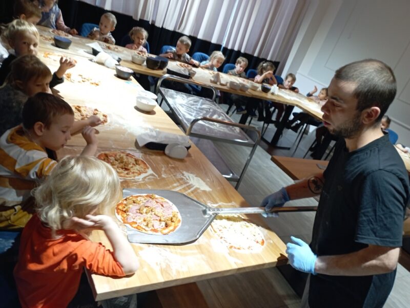 Na zdjęciu pizzerman bierze od dzieci przygotowane pizze, w celu włożenia ich do pieca.