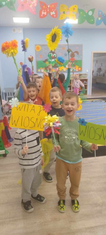 djęcie pierwsze przedstawia dzieci ustawione w rzędzie. Dzieci trzymają w dłoniach tabliczki z napisami: " Wiosna" i " Witamy wiosnę" oraz kwiaty wykonane z papieru i krepy.