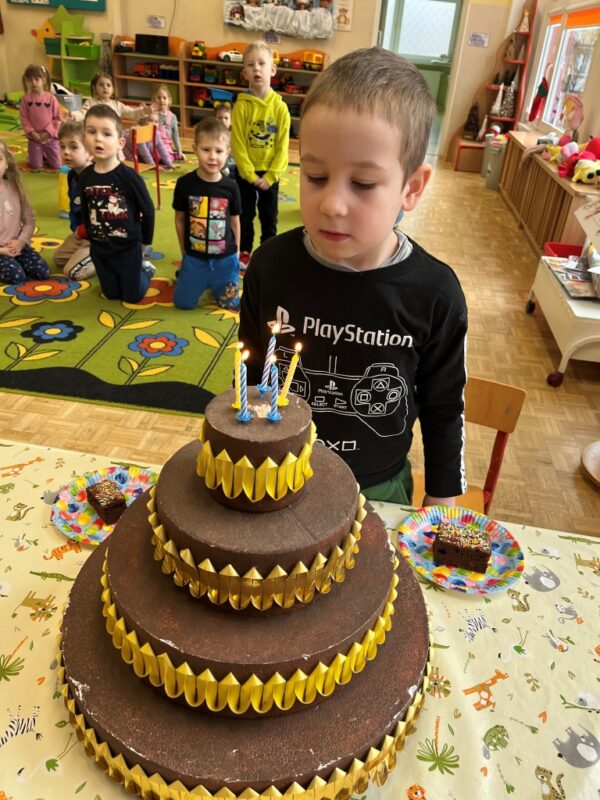Na zdjęciu przedstawiony jest Grzesiu, który po pomyśleniu życzenia dmucha pięć kolorowych, urodzinowych świeczek. Obok niego stoi kawałek słodkiego, czekoladowego tortu, który wykonał wspólnie z mamą.
