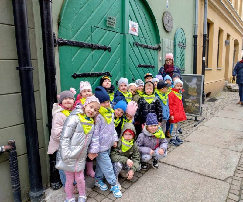 Zdjęcie pierwsze przedstawia grupę dzieci wraz z nauczycielem na tle wejścia do Muzeum.
