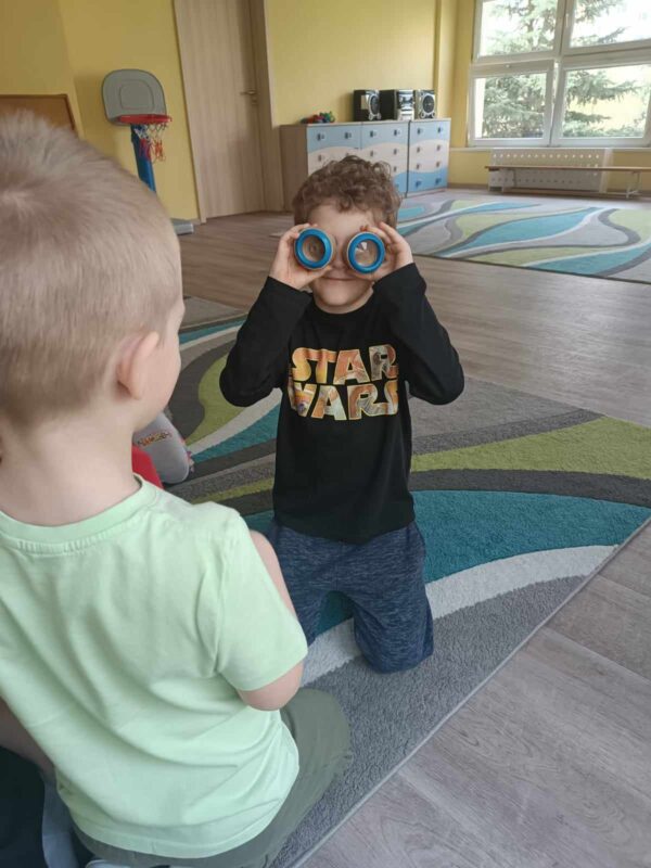 Zdjęcie pierwsze przedstawia chłopca, który przykłada sobie do oczu urządzenie wyglądem przypominające lornetkę, dzięki któremu może oglądać świat z perspektywy mrówki. Inny chłopiec uważnie mu się przygląda.