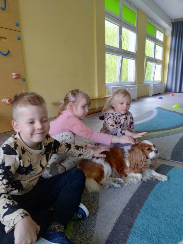 Zdjęcie pierwsze przedstawia troje dzieci- dwie dziewczynki i chłopca siedzących na dywanie i głaszczących psa.