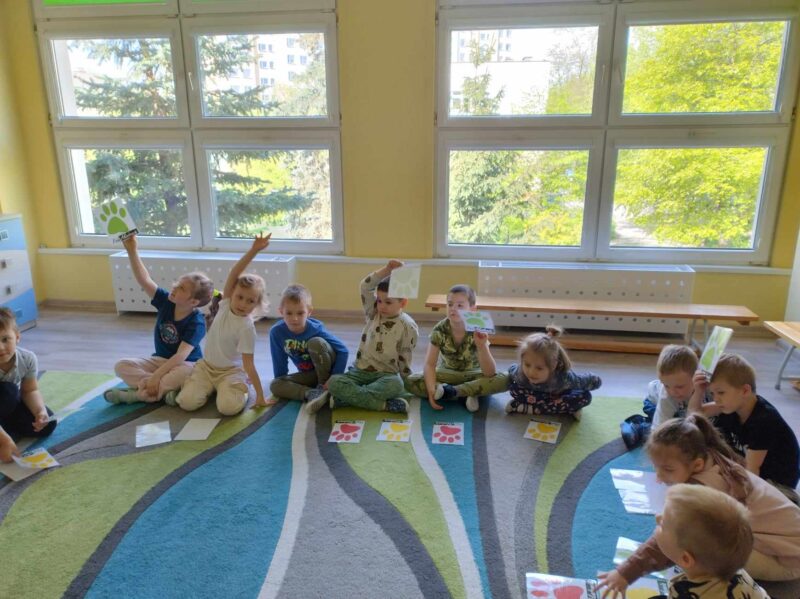 Zdjęcie drugie przedstawia grupę dzieci siedzącą na dywanie. Dzieci podnoszą w górę karty przedstawiające psie łapy w różnych kolorach.