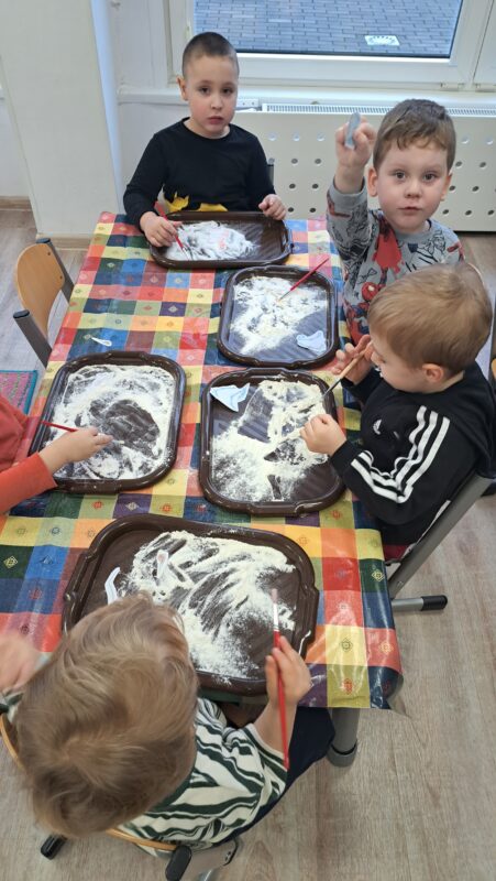 Dzieci siedzą przy stolikach, na których, na tackach pod mąką ukryte są papierowe szkielety. Dzieci pędzlami odkrywają szkielety dinozaurów.