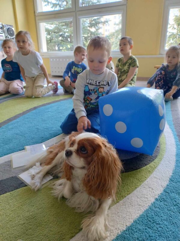 Na pierwszym planie widoczny jest chłopiec siedzący na dywanie i głaszczący psa. Obok chłopca znajduje się duża nadmuchana kostka do gry.  W tle widać inne dzieci siedzące na dywanie.