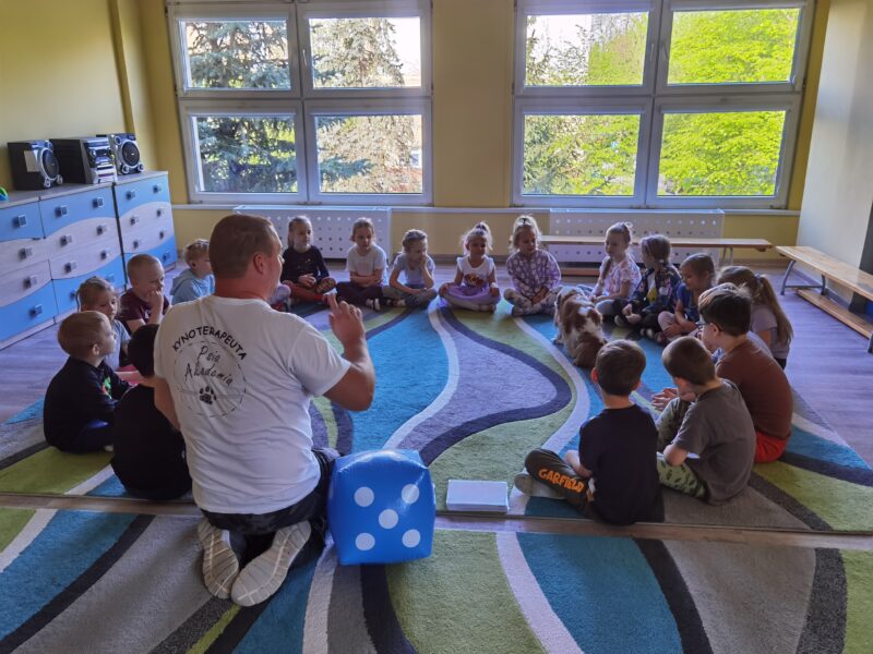 Dzieci siedzą w kole na dywanie, widać pomoce dydaktyczne czyli kostkę, dzieci uwaznie sluchaja prowadzącego