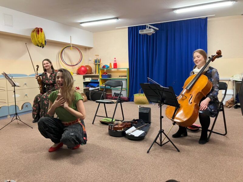 Zdjęcie przedstawia panie prowadzące spotkanie z dziećmi. Jedna ma wiolenczelę, druga skrzypce, a trzecia przedstawia dzieciom instrumenty.