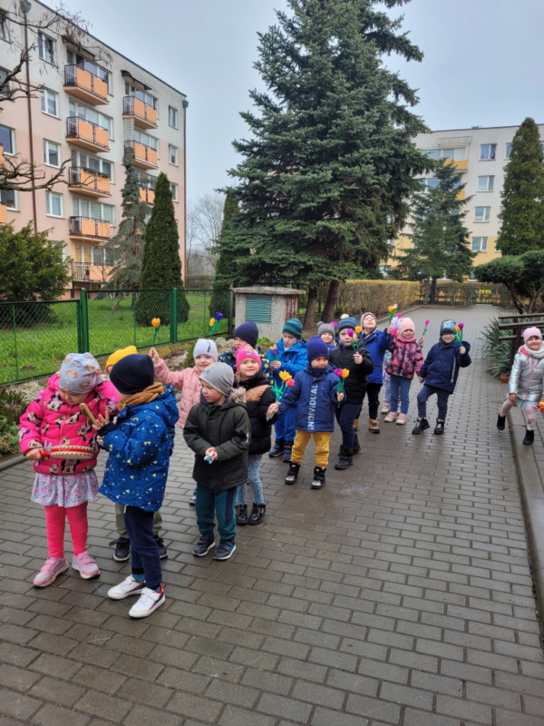 Dzieci ustawione parami przed budynkiem przedszkola, przygotowane do marszu żegnającego zimę. Dzieci w rękach trzymaja kwaity z krepy bądź instrumenty.