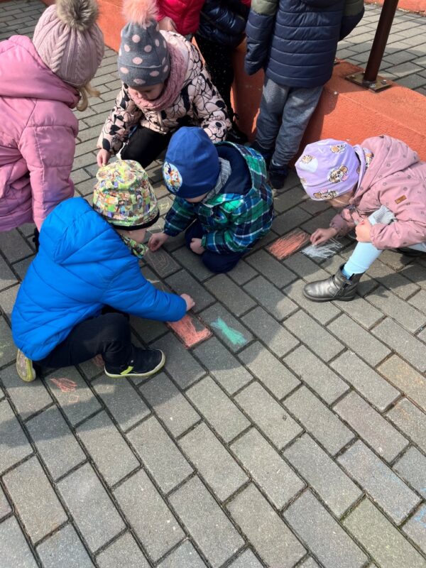 Dzieci ze skupieniem oraz zadowoleniem kolorują kredą prostokąty na przedszkolnym chodniku, aby kolorami przywołać szybciej ciepłą wiosnę.