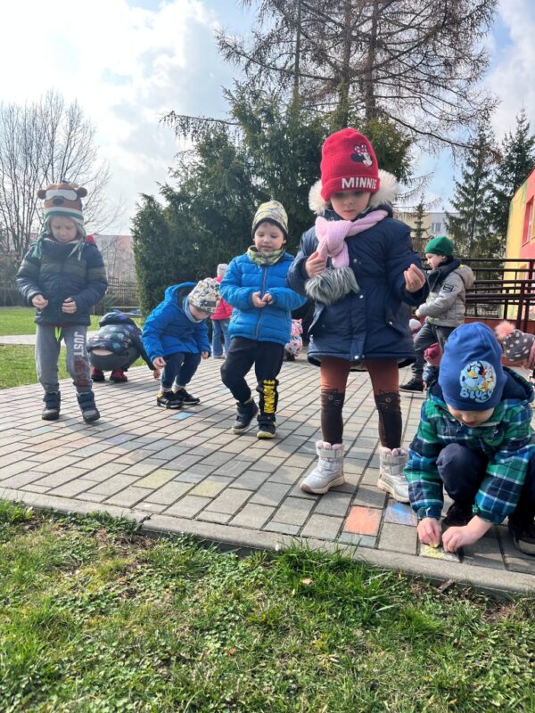 Dzieci ze skupieniem oraz zadowoleniem kolorują kredą prostokąty na przedszkolnym chodniku, aby kolorami przywołać szybciej ciepłą wiosnę.