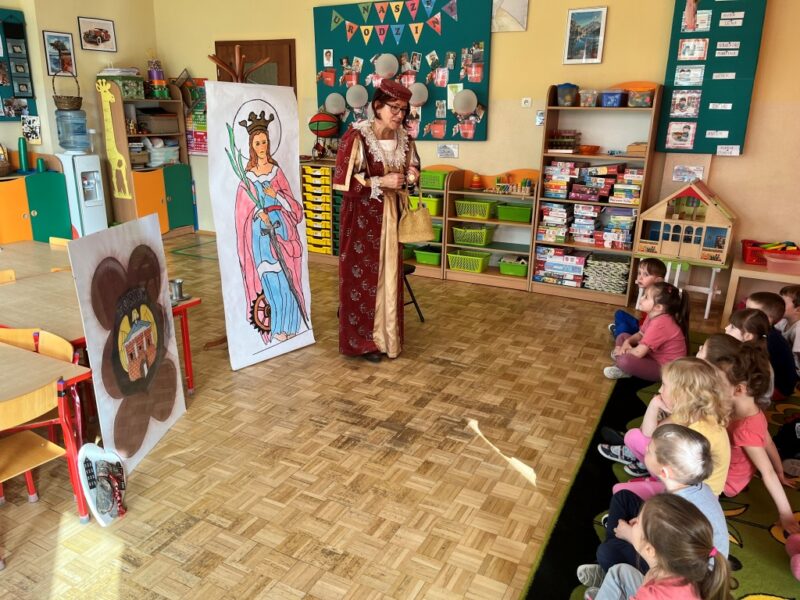Dzieci siedzą na dywanie i z uwagą oraz zainteresowaniem słuchają opowieści (przebranej w strój danej księżniczki) Pani Przewodnik na temat historii naszego miasta - Torunia. 