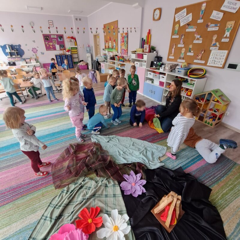 Na zdjęciu dzieci  siedzą na dywanie Sali przedszkolnej, na której znajdują się kwiaty z krepy, sztuczne ognisko.