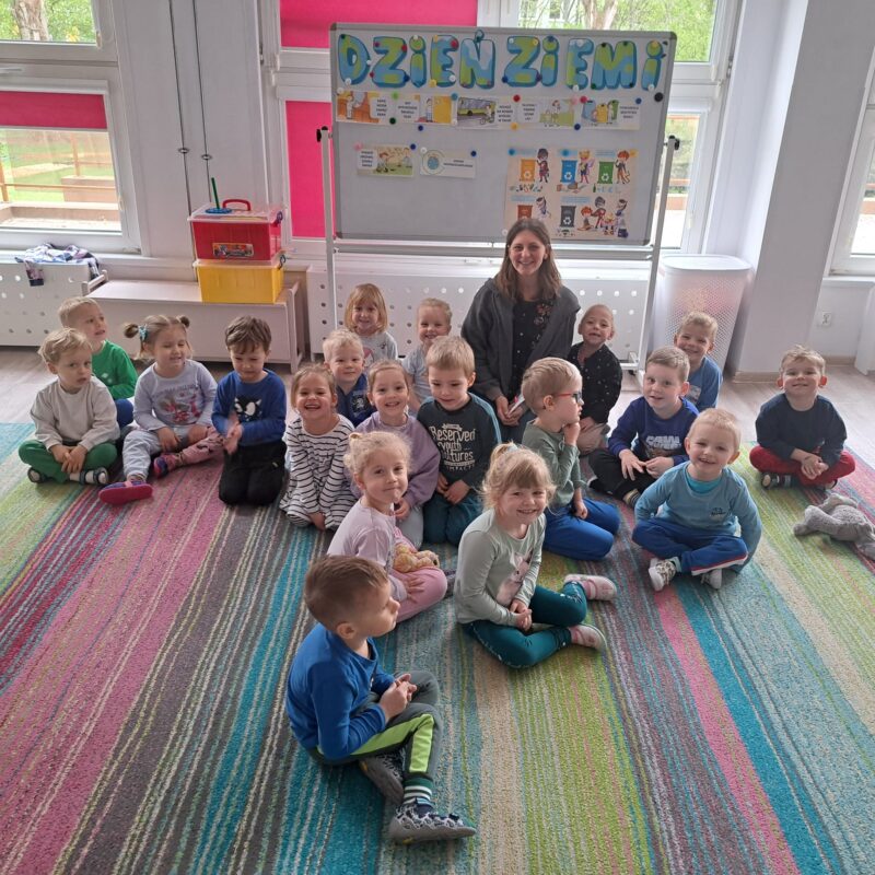 Fotografia przedstawia dzieci bawiące się w sali przedszkolnej na dywanie. Dzieci chowają się na dywanie w pozycji skulonej, a nauczyciel przykrywa je chustą animacyjną.