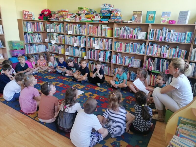 Dzieci siedzą skrzyżnie w kręgu na dywanie. Za nimi regały z książkami. Pani bibliotekarka siedzi z dziećmi w kręgu i czyta wiersz. 