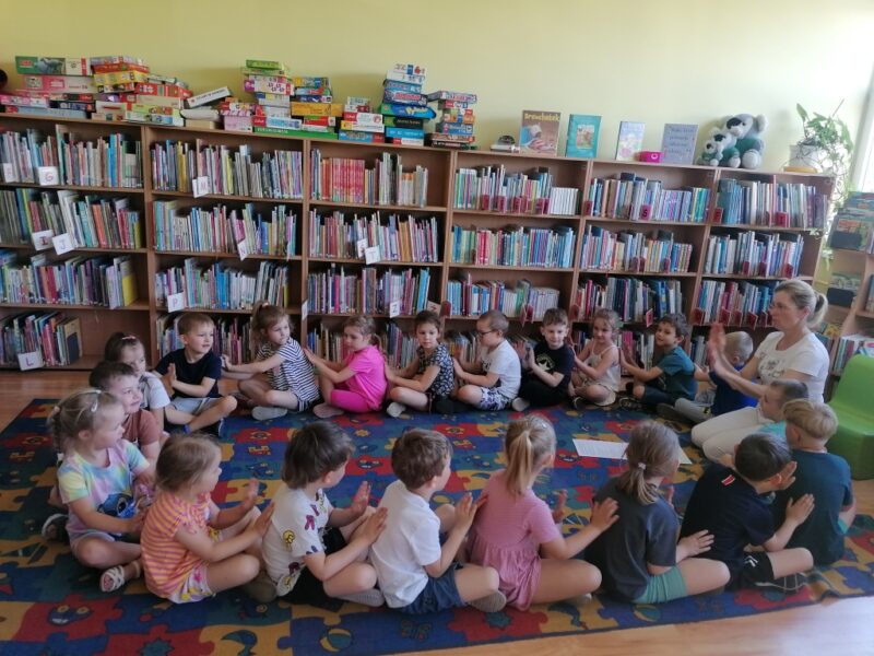 Dzieci siedzą skrzyżnie w kręgu na dywanie bokiem do środka koła i wykonują masaż obiema rękami rówieśnikowi przed sobą. Pani bibliotekarka czyta im wiersz i pokazuje ruch do masażyku.