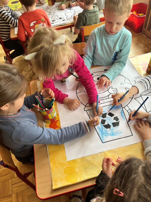 Dzieci grupowo siedzą przy stolikach i w skupieniu oraz z radością wykonują swoją pracę plastyczną. Kolorują kolorowymi kredkami dużą kolorowankę ukazującą eko Ziemię. 
