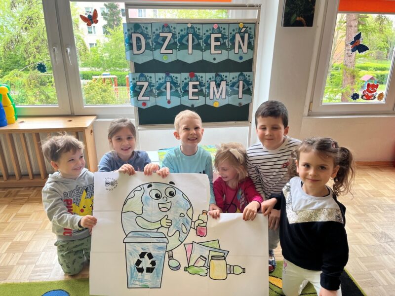 Dzieci w swojej grupie z szerokimi uśmiechami prezentują wykonaną i odpowiednio pokolorowaną przez siebie eko planetę Ziemię, która segreguje odpady. Za nimi, w tle, na zielonej tablicy znajduje się napis ''Dzień Ziemi''. 