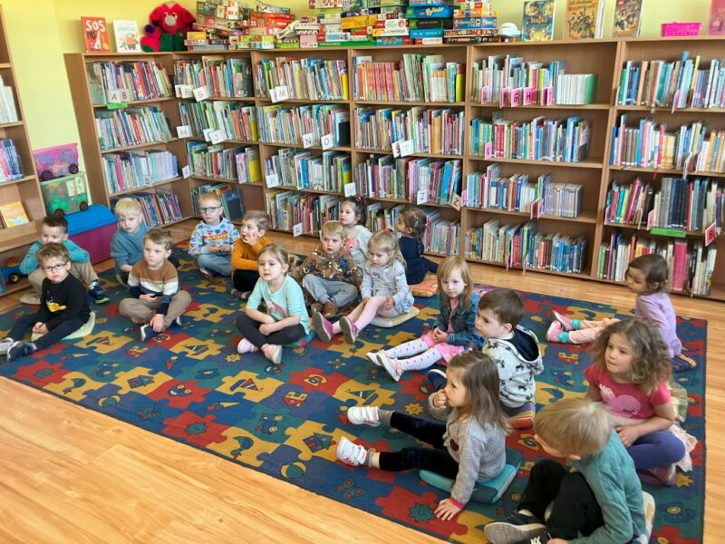 Dzieci siedzą na dywanie na miękkich poduchach. Wokół nich znjdują się reagały z książkami dla dzieci.
