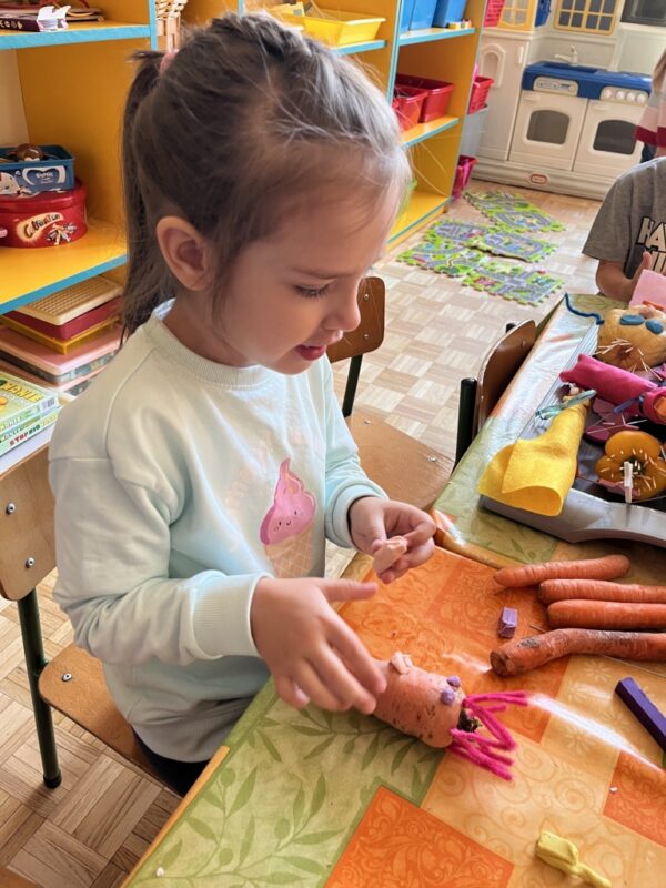 Dziewczynka z wielką dokładnością i uwagą przykleja z beżowej plasteliny nos do swojej leżącej na stoliku marchewki. Ma ona już przyklejone fioletowe oczy oraz beżowy, szeroki uśmiech - również są one zrobione z plasteliny. 