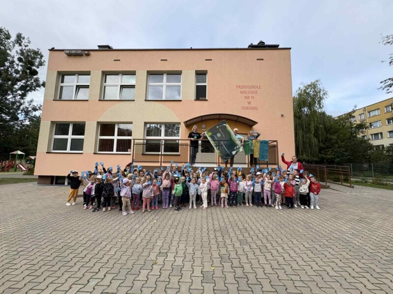Dzieci stoją na kostce brukowej na tle przedszkola. Mają ubrane rękawiczki niebieskie jednorazowe na jednej ręce, uśmiechają się do zdjęcia, rekę z rękawiczką mają w górze. Na podwyższeniu stoją 3 nauczycielki z flagą akcji „sprzątania świata” i workami do segregacji śmieci.