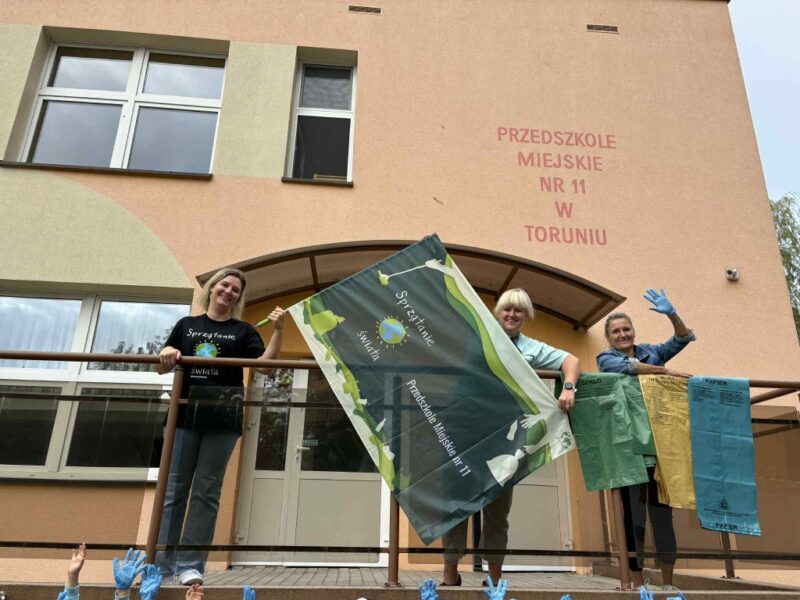 Nauczycielki stoją na tle budynku przedszkola ,za barierką. Nauczycielka po lewej ma ubraną koszulkę akcji „Sprzątania świata „ i trzyma w rękach kij od flagi związanej z akcją. Nauczycielka na środku trzyma flagę za materiał. Nauczycielka po prawej trzyma kolorowe worki do segregacji odpadów i macha do zdjęcia. Wszystkie się uśmiechają.